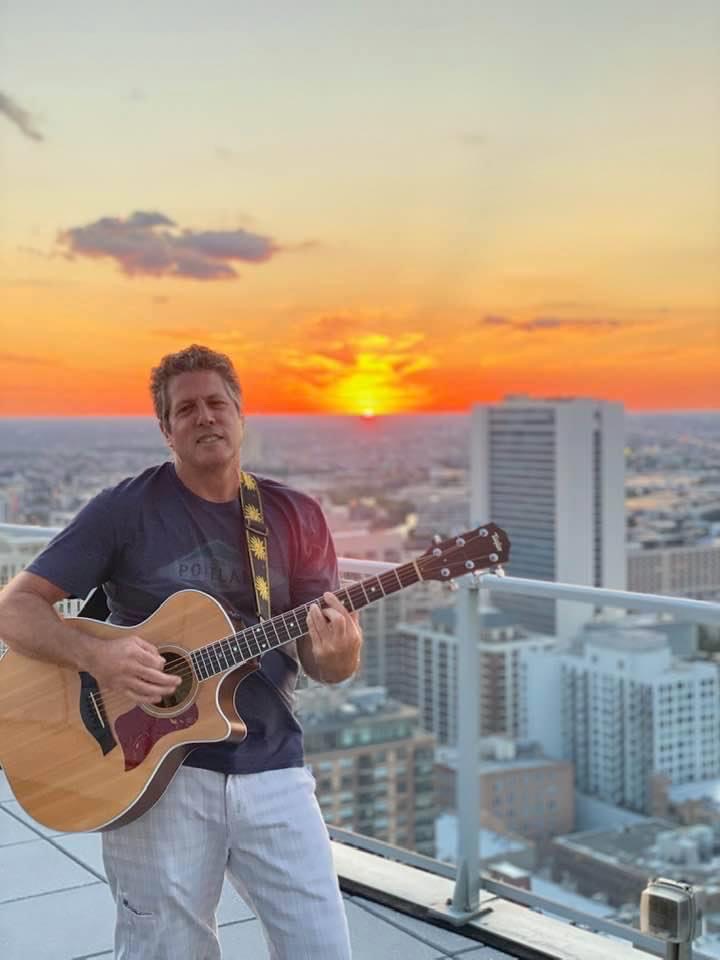 Ron Cohler playing acoustic guitar on a Chicago rooftop at sunset